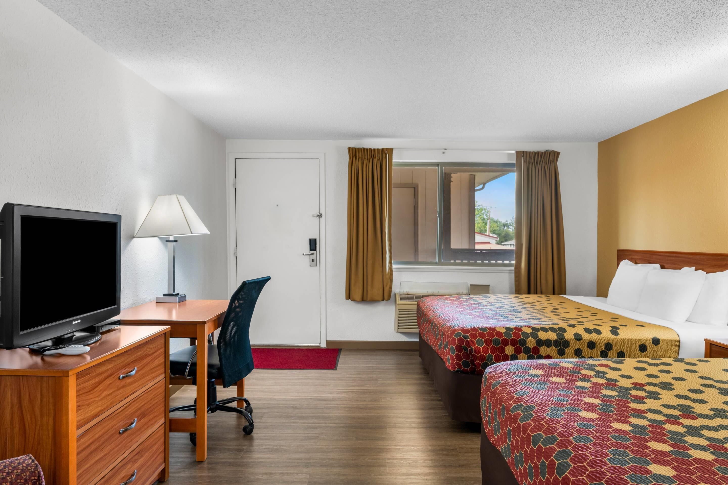 A hotel room with a desk, television, two beds, a chair, and a window with curtains.