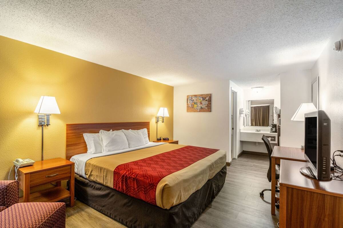 A hotel room with a bed, bedside tables, a desk, a chair, a television, a mirror, and a bathroom visible through an open door.