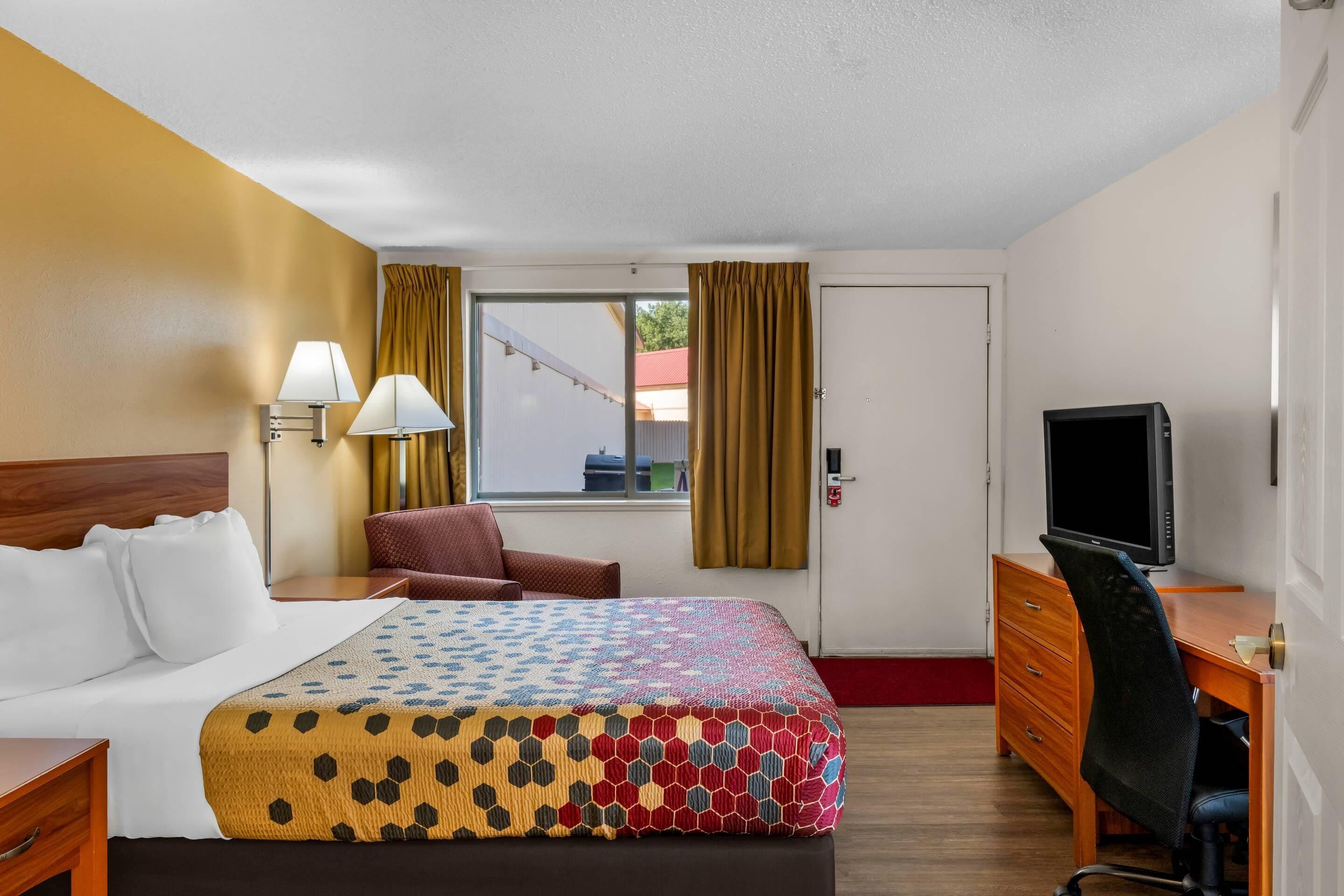 Habitación de hotel con una cama, un escritorio con computadora, una silla, televisor y ventana con cortinas.
