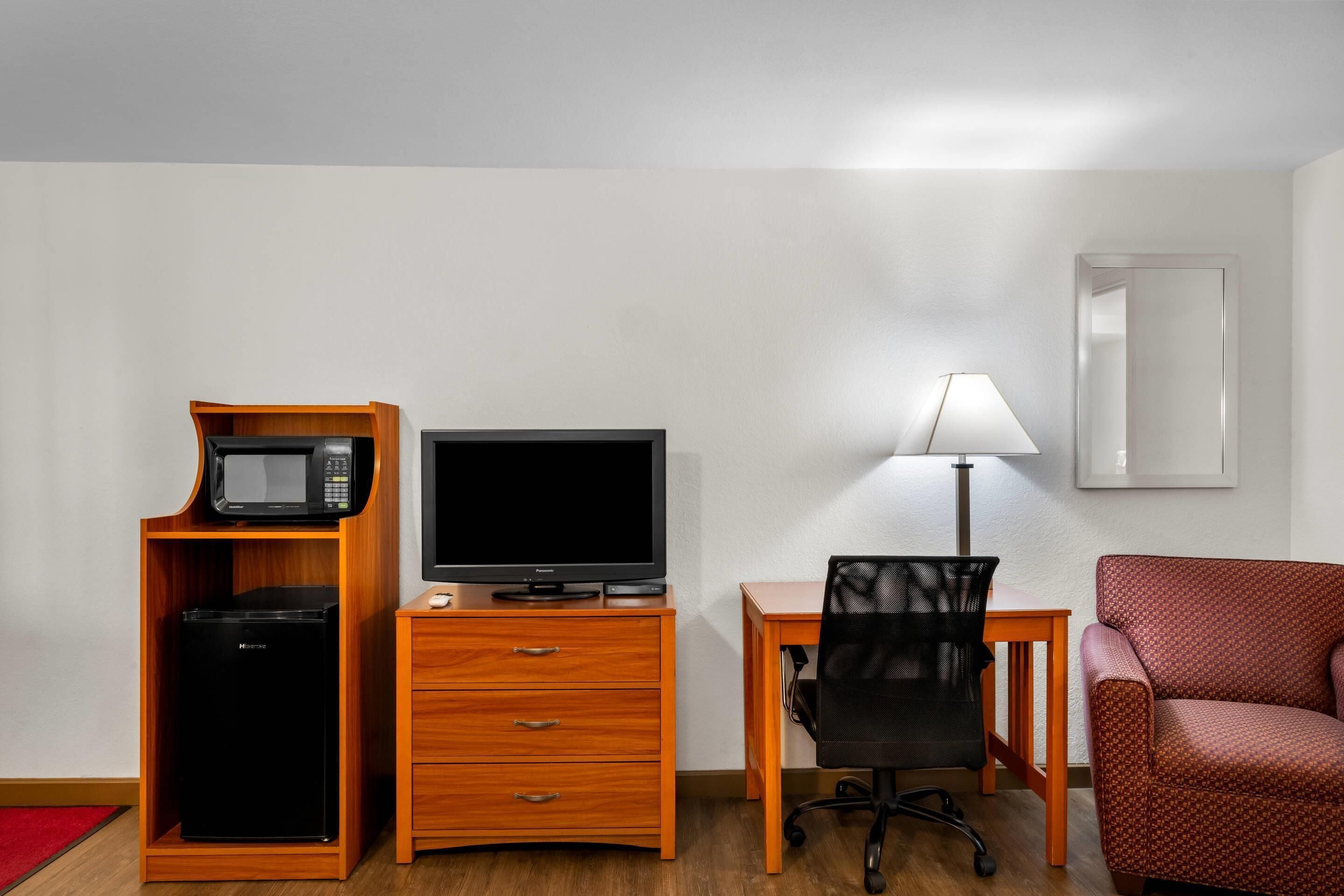 Une chambre d’hôtel équipée d’un bureau en bois, d’une télévision, d’un réfrigérateur, d’une chaise et d’une lampe.