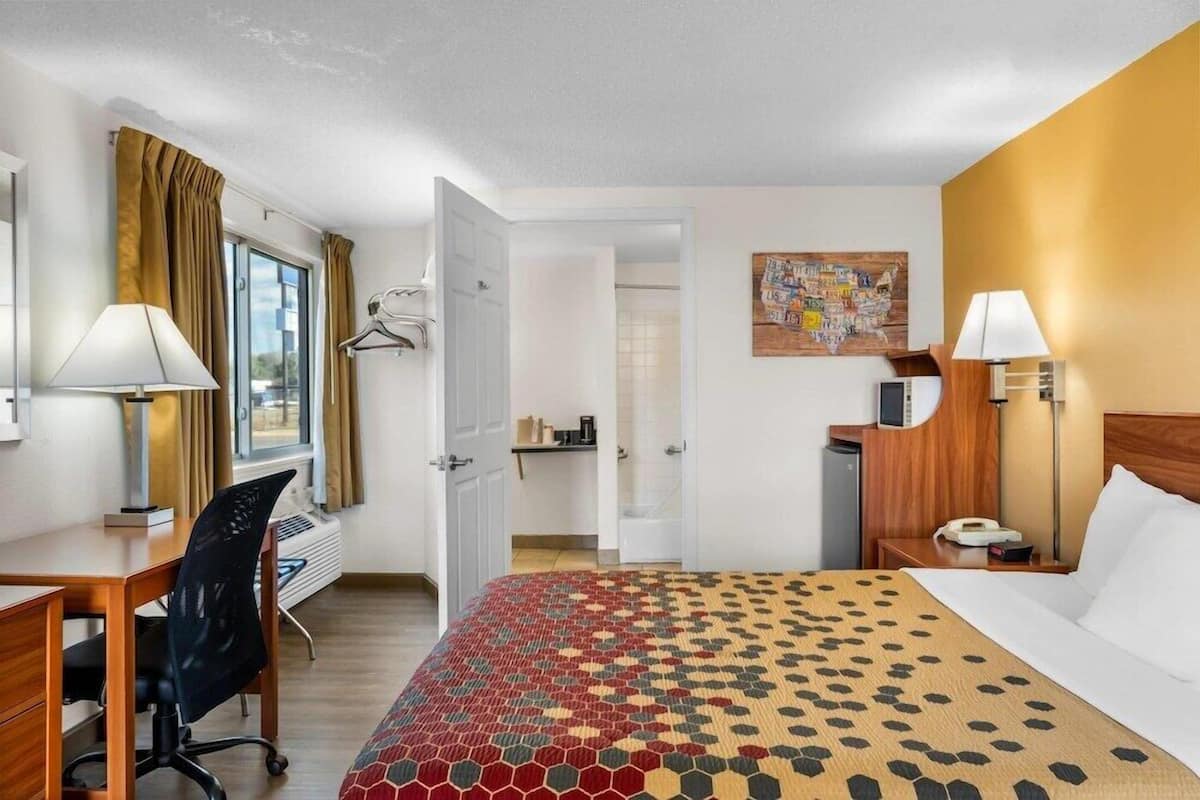 A hotel room with a bed, desk, chair, lamp, and a painting on the wall.