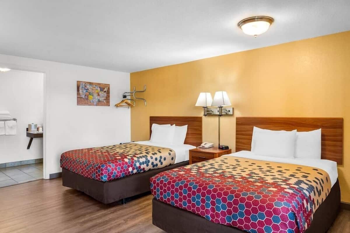 A hotel room with two beds, a nightstand with a lamp, a wall-mounted coat rack, and a painting.