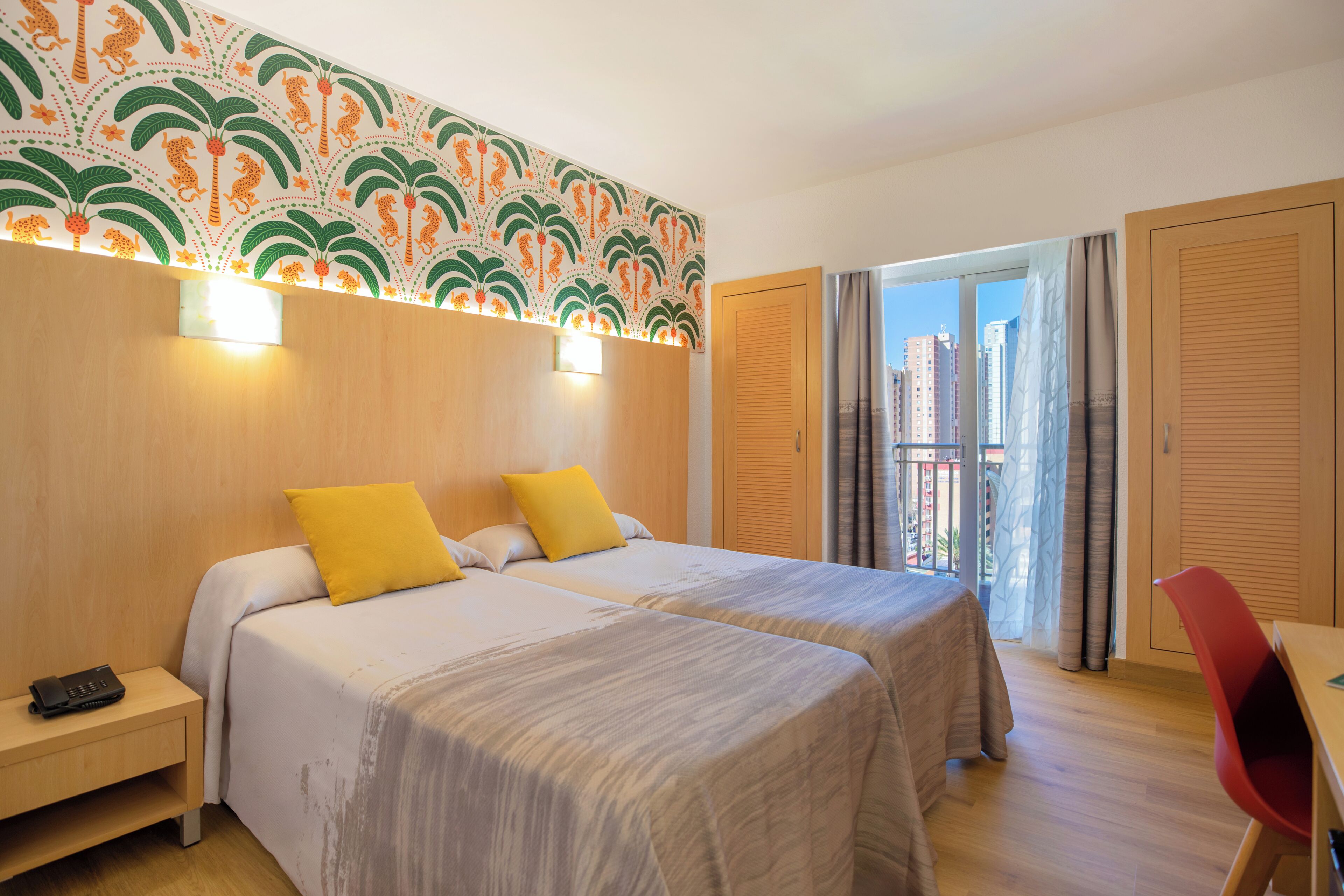 Habitación de hotel con dos camas, una mesita de noche, un escritorio y un balcón con vistas a edificios.