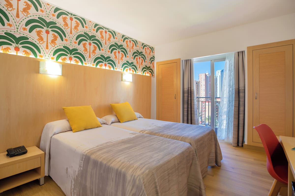 Habitación de hotel con dos camas, una mesita de noche, un escritorio y un balcón con vistas a edificios.