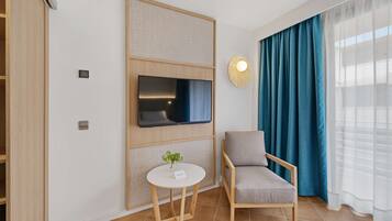 Una habitación moderna con un televisor de pantalla plana montado en un panel de madera, una pequeña mesa redonda con una tarjeta de nombre, un sillón tapizado gris y un gran ventanal con cortinas azules.