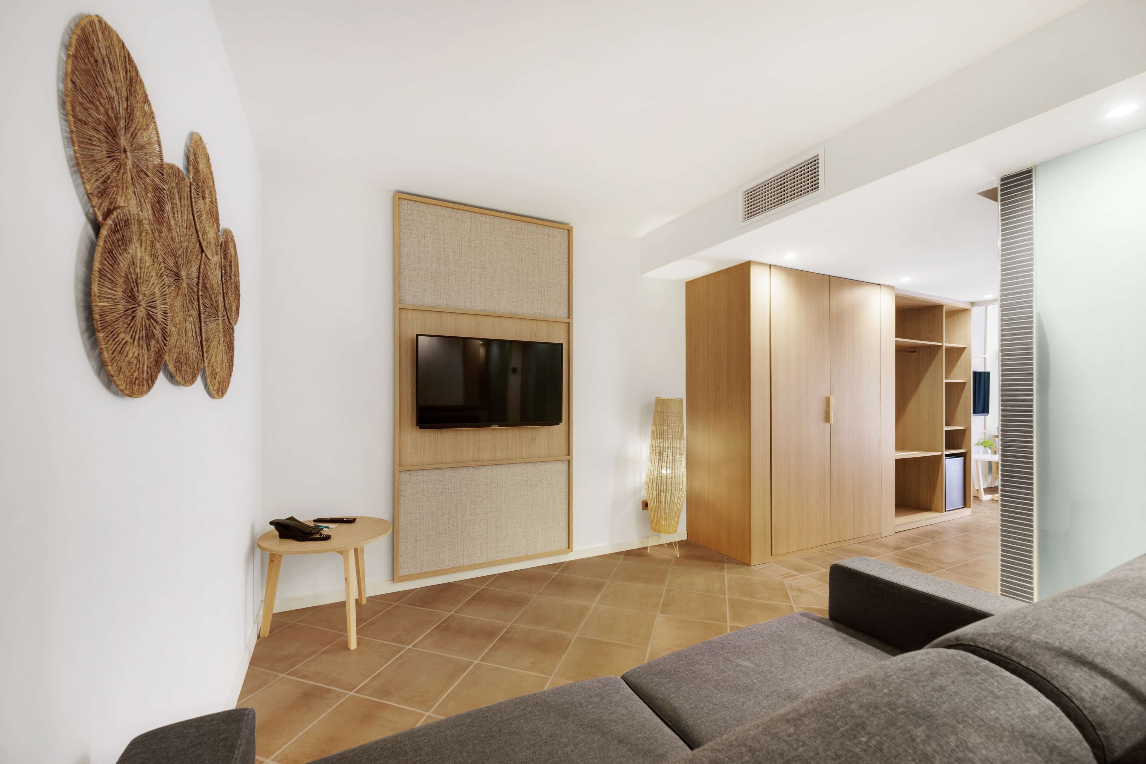 A modern living room with a grey sofa, a flat-screen TV mounted on a wooden panel, a small wooden table with a chair, and a large woven wall art piece.