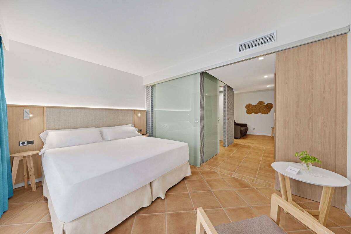 A modern hotel room with a large bed, a small table with a plant, and a bathroom visible through a glass door.