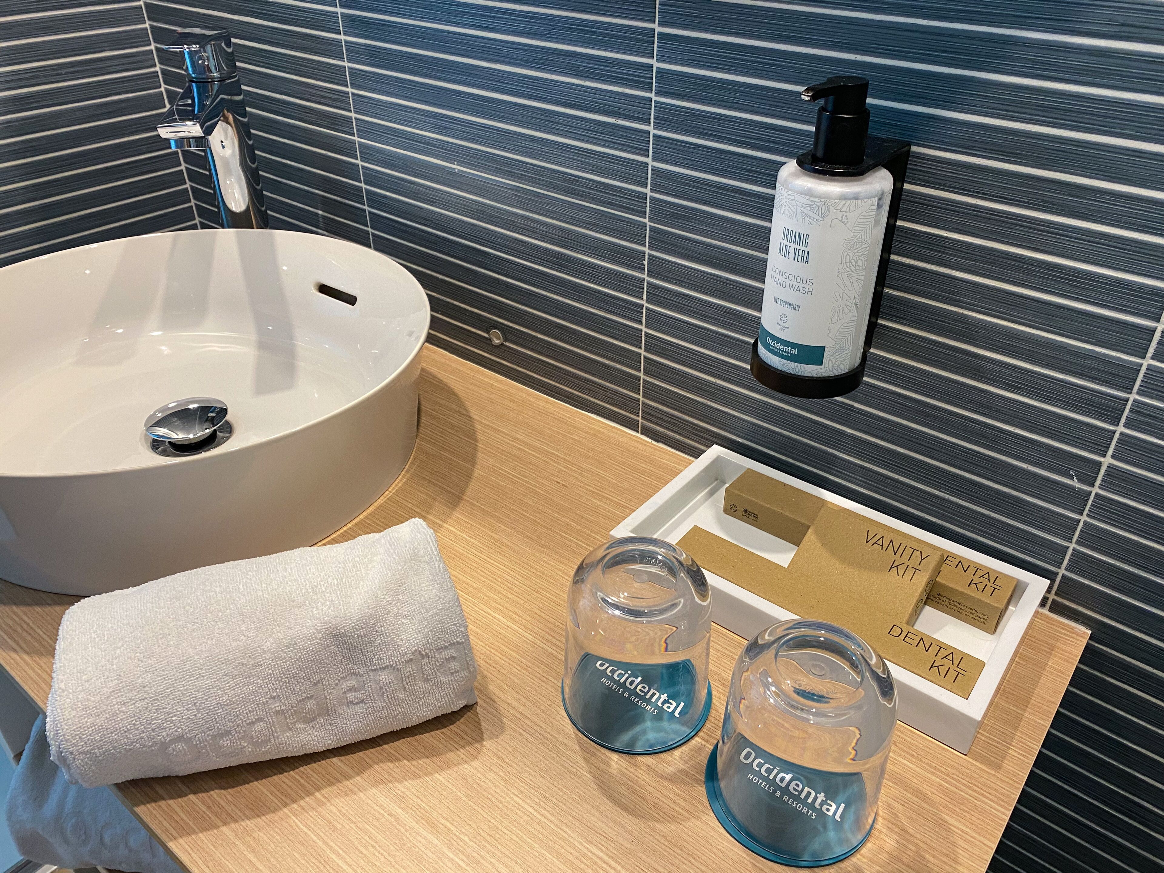 A bathroom with a white sink, a bottle of organic aloe vera hand wash, two glasses of water, a towel, and a vanity kit.