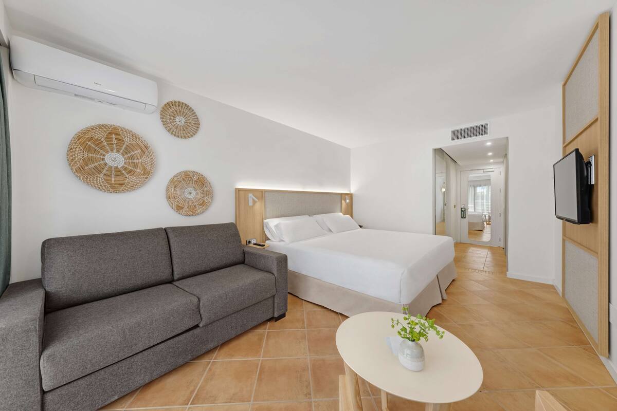 A modern hotel room with a grey sofa, a large bed, a small round table with a plant, and decorative wall art.