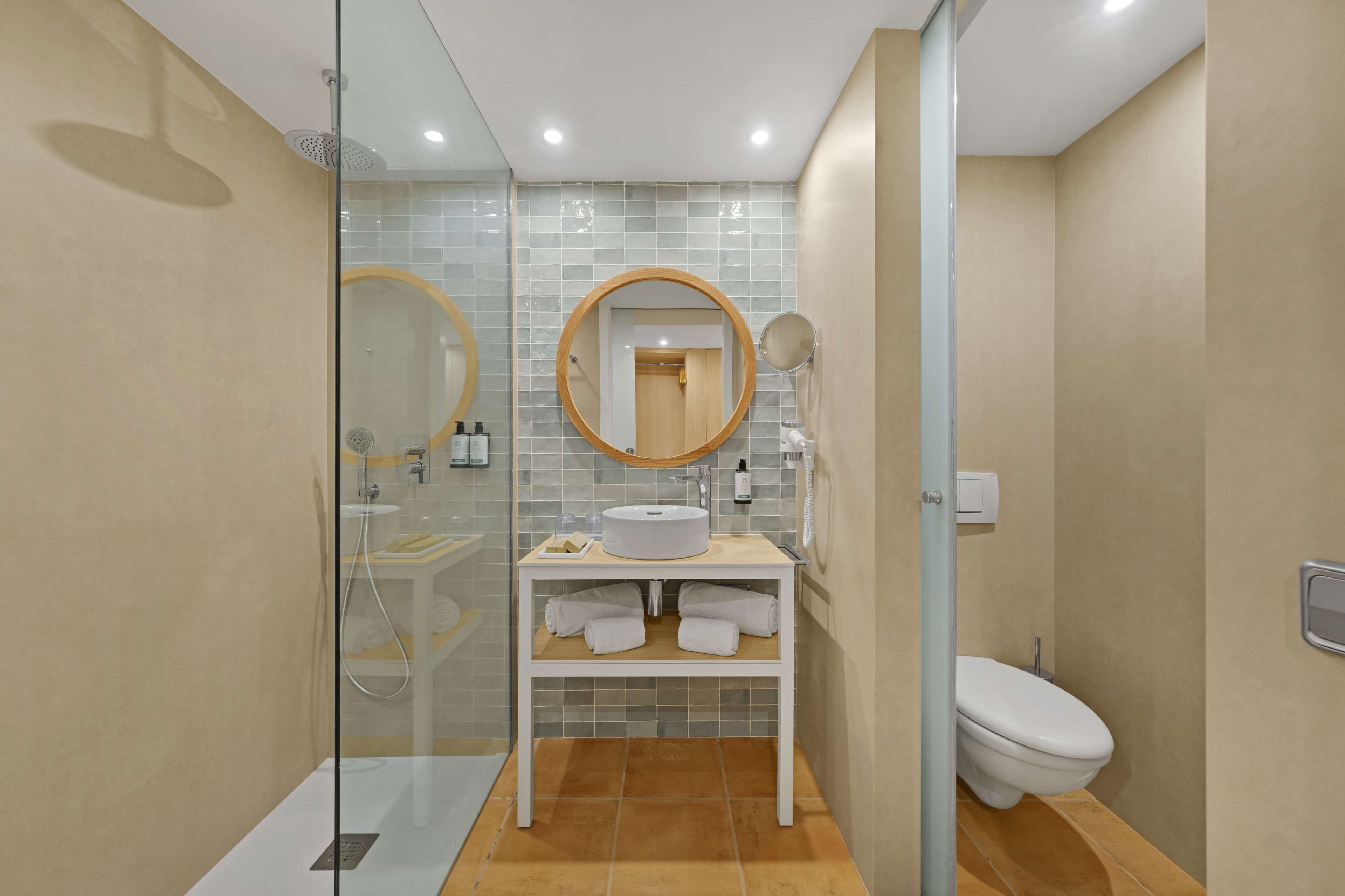 A modern bathroom with a glass-enclosed shower, a round mirror, a sink with a towel rack, and a toilet.