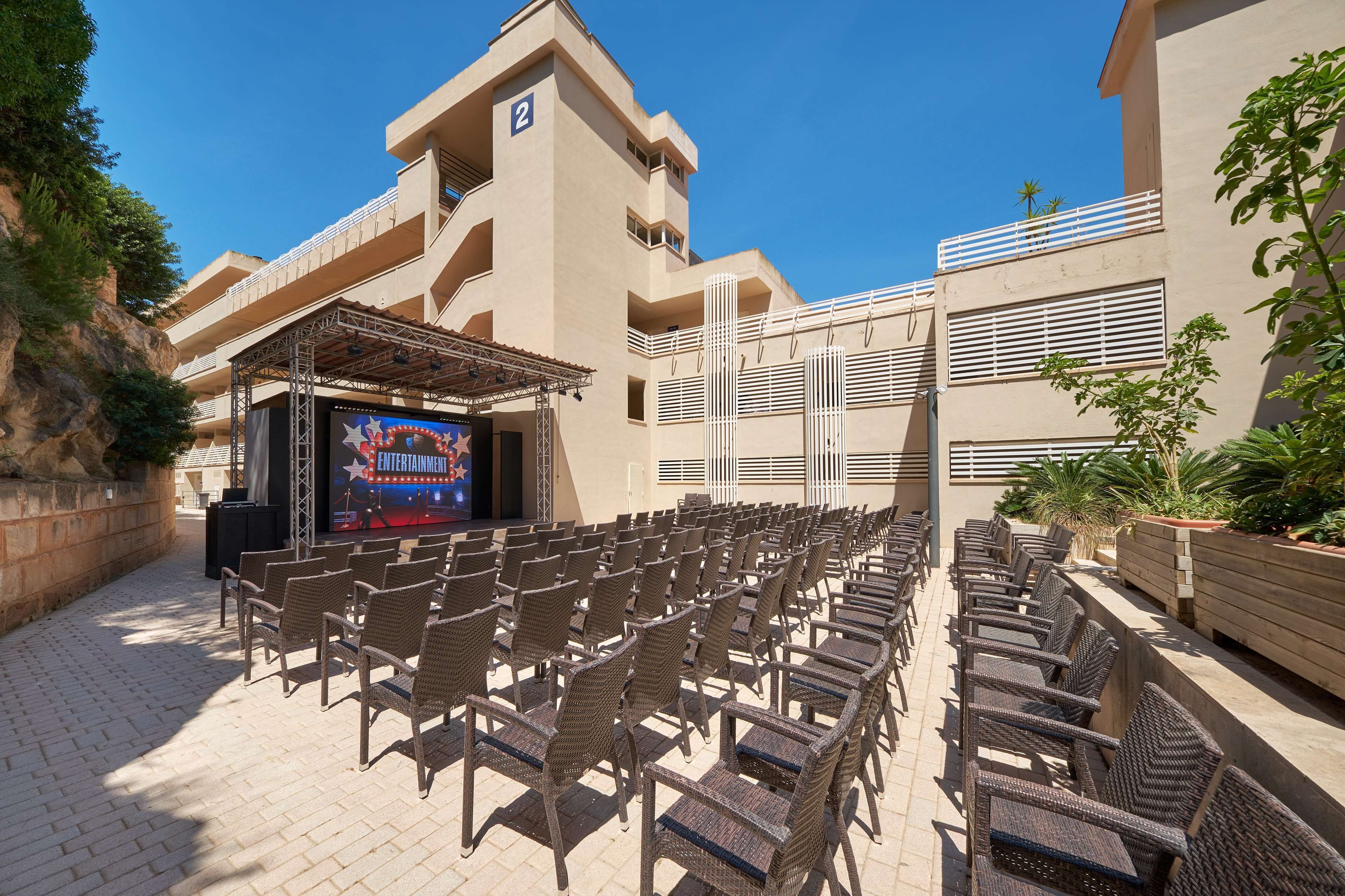 An outdoor seating area with wicker chairs arranged in rows, a stage with a large screen, and a building with balconies and a number '2' sign.