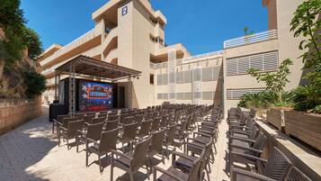 An outdoor seating area with wicker chairs arranged in rows, a stage with a large screen, and a building with balconies and a number '2' sign.