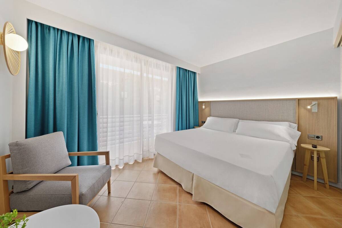 A hotel room with a bed, a chair, a small table, and a wall-mounted light.