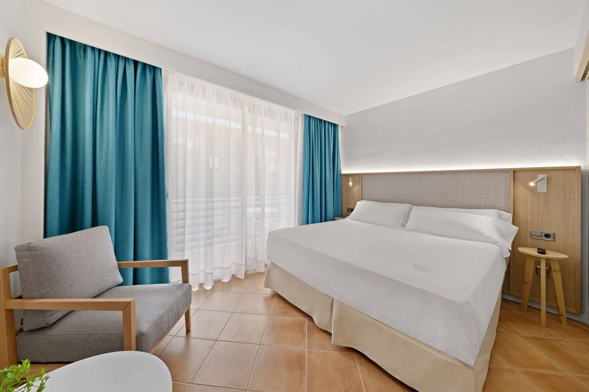 A hotel room with a bed, a chair, a bedside table, and a wall-mounted lamp.
