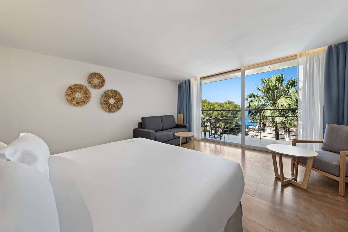 A hotel room with a large bed, a sofa, a small table, and a balcony with a view of palm trees.