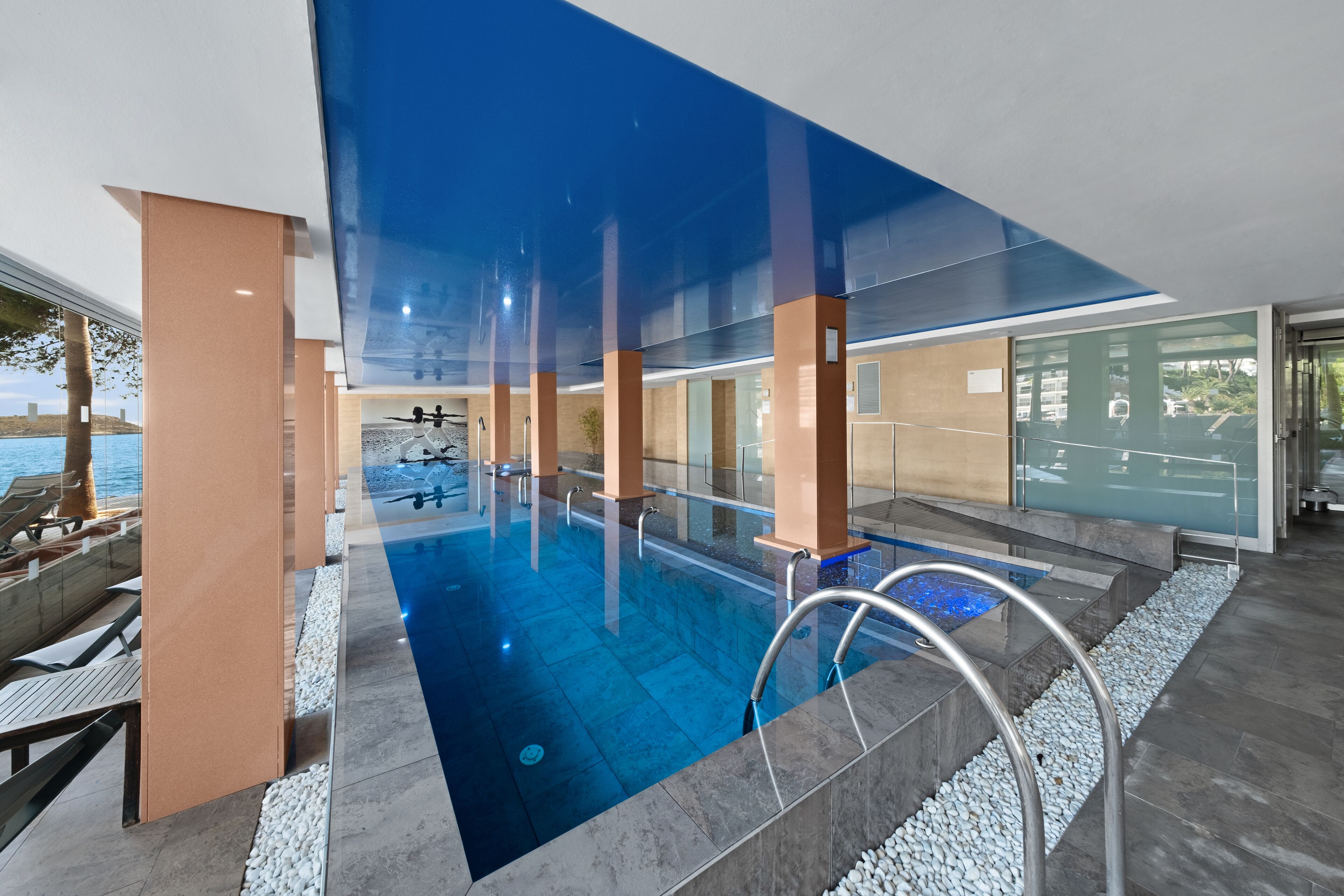 Una piscina interior moderna con paredes de vidrio transparente, vista al mar y una terraza con sillones de descanso.