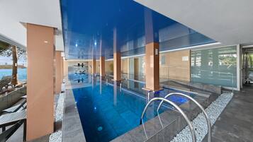 Una piscina interior moderna con paredes de vidrio transparente, vista al mar y una terraza con sillones de descanso.