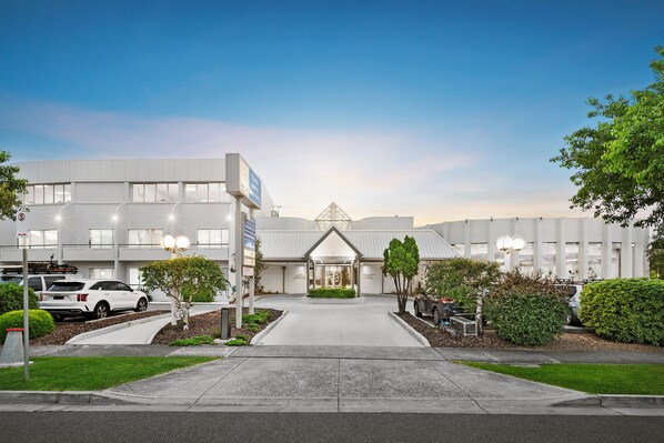 Un edificio moderno con entrada central, zona de estacionamiento y jardines bien cuidados.