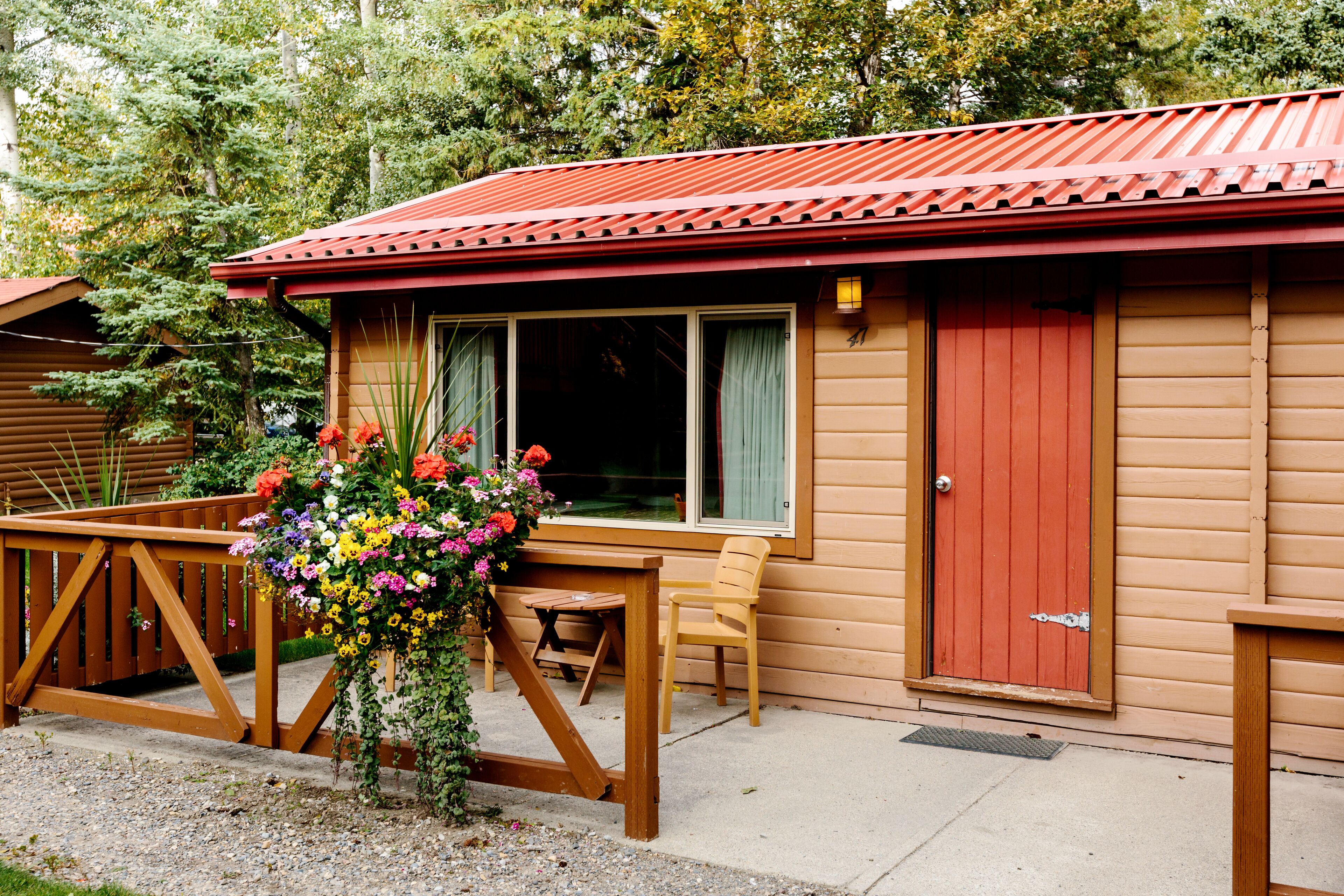 Una cabana de fusta amb una porta vermella, una finestra amb cortines, un balcó de fusta i un arranjament floral.