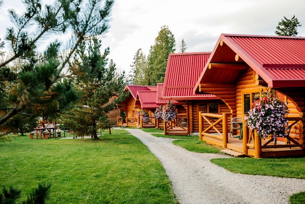 A row of log cabins with red roofs, a gravel path, and a picnic table in a grassy area.