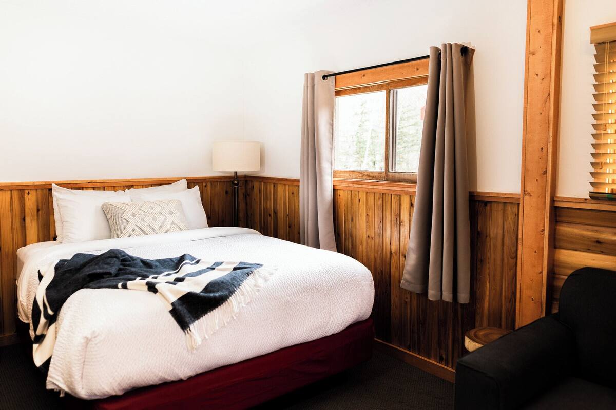 Un dormitorio con cabecera de madera, cama con ropa de cama blanca y un toque rojo, una lámpara de noche, una ventana con cortinas y una silla.
