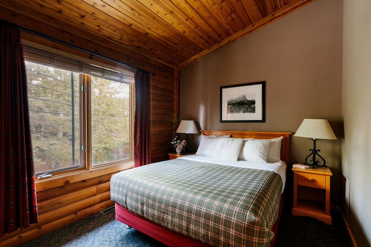 Un dormitorio de cabaña de madera con una cama grande, dos mesitas de noche con lámparas, una ventana con cortinas y un cuadro en la pared.