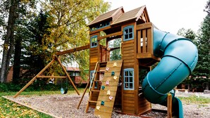 A wooden play structure with a slide and climbing wall, surrounded by trees and a grassy area. - Miette Mountain Cabins (Pocahontas)