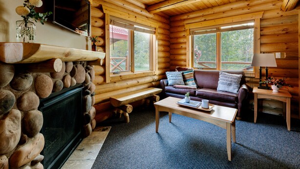 A log cabin interior with a stone fireplace, a leather sofa, a wooden coffee table, and large windows. - Miette Mountain Cabins (Pocahontas)