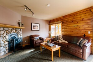 A cozy living room with a stone fireplace, leather sofas, a wooden coffee table, and a large window with wooden shutters. - Miette Mountain Cabins (Pocahontas)