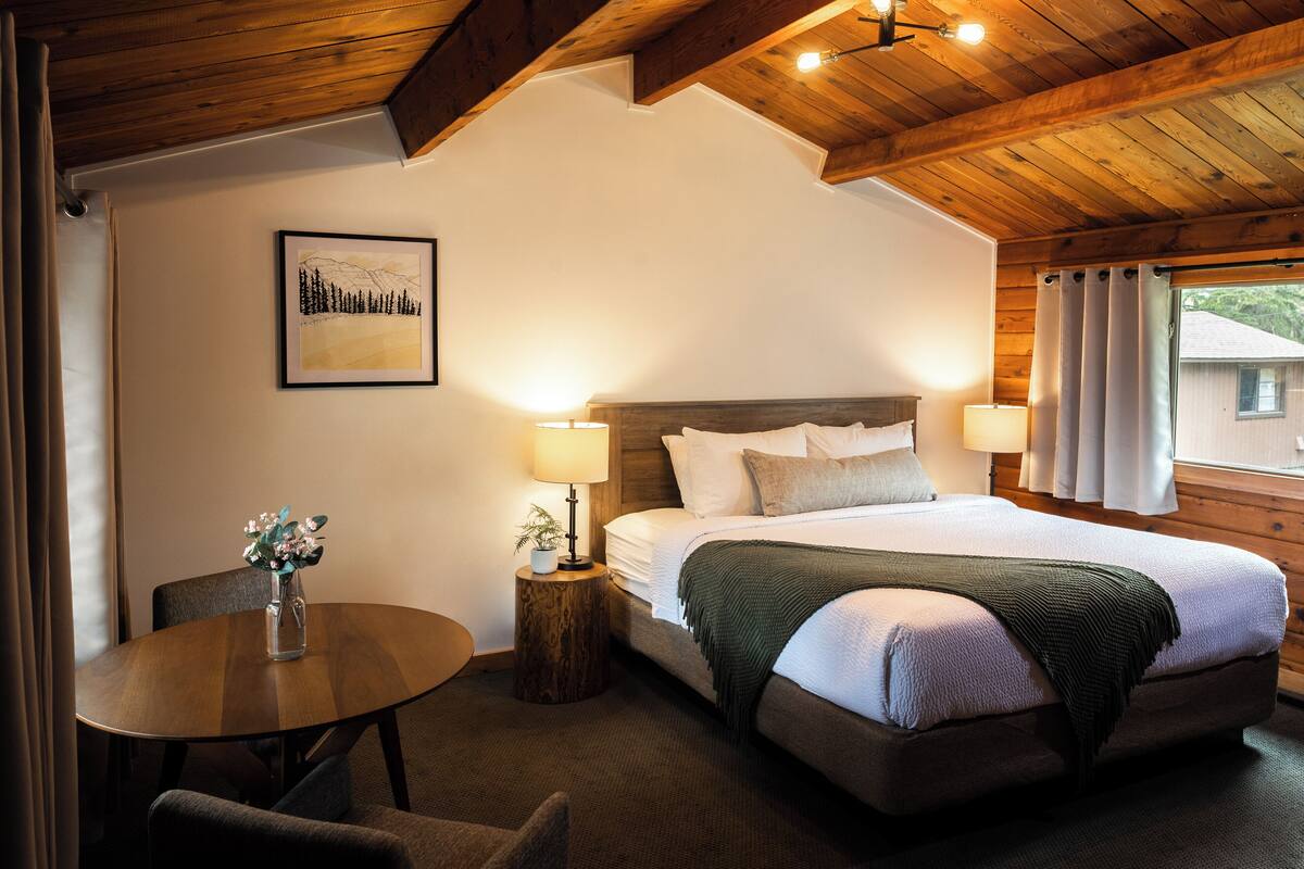 Un dormitorio con una cama grande, lámparas de noche, una mesita redonda con un florero y un cuadro en la pared.