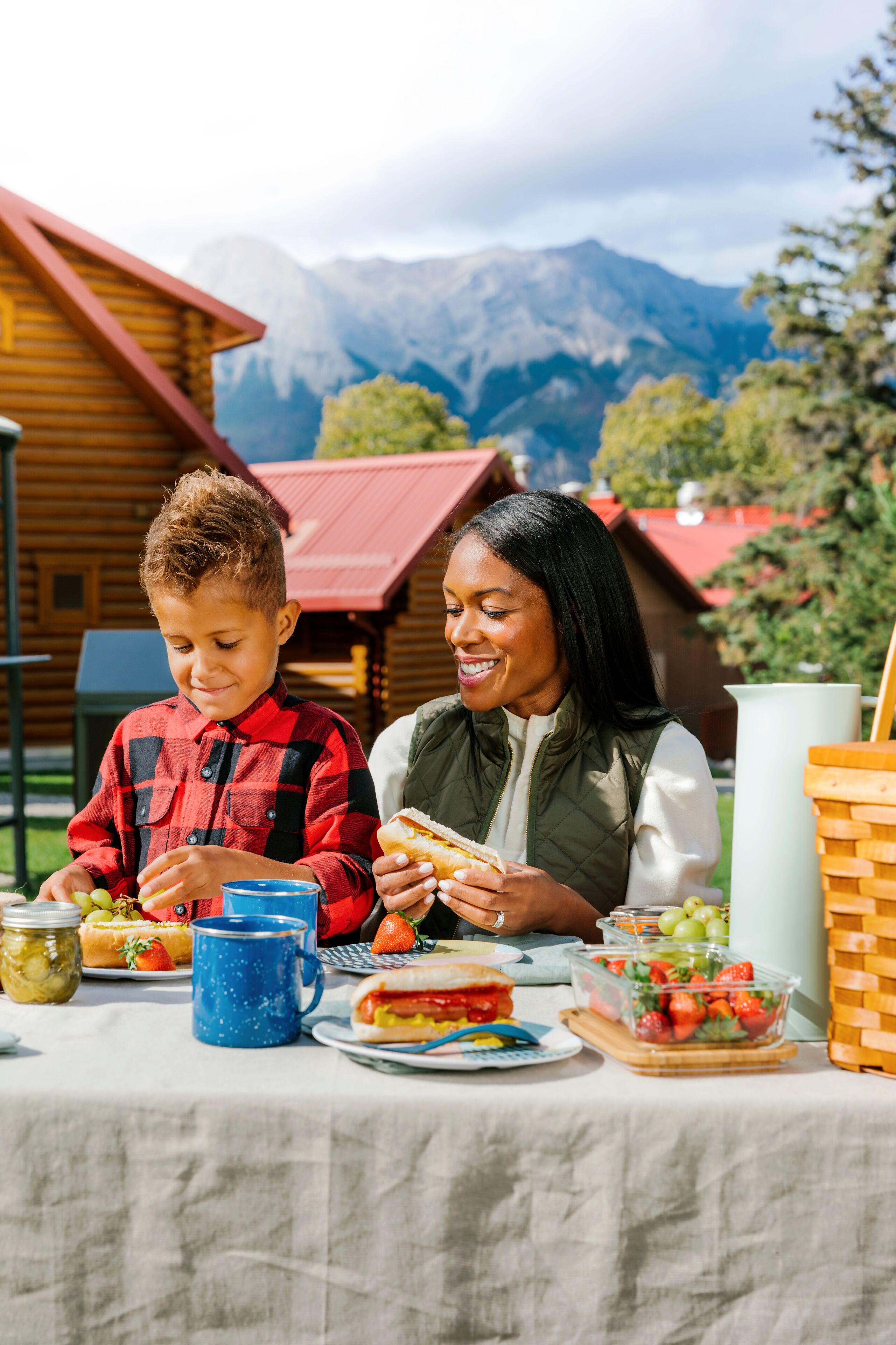 A dining table with food and drinks, a wooden cabin in the background, and mountains in the distance.