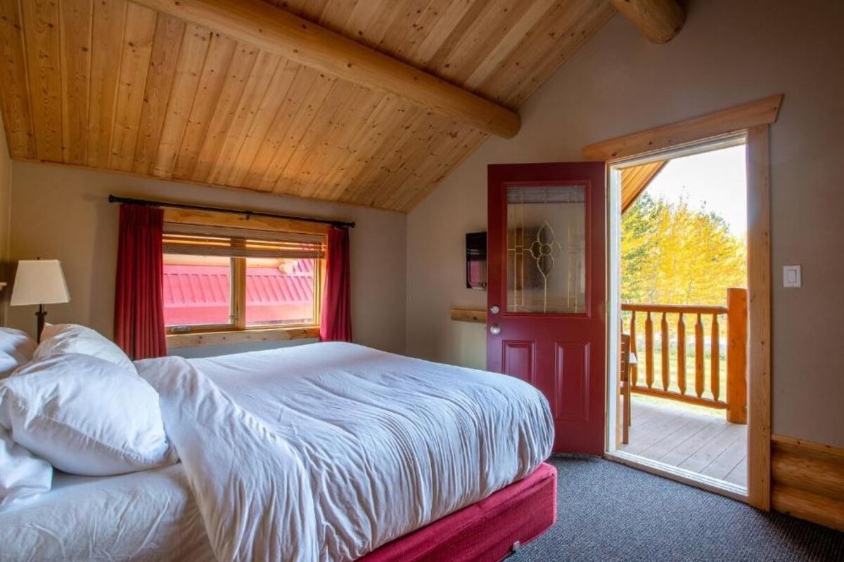 Un dormitorio con techo de madera, cama con ropa blanca, puerta roja y balcón con barandilla de madera.