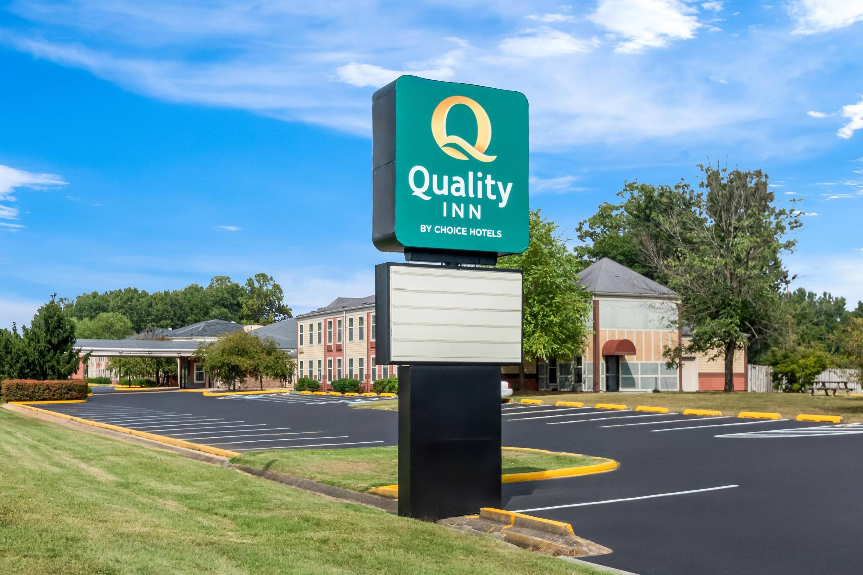 Skilt for Quality Inn med Choice Hotels logo, parkeringsplads og bygninger i baggrunden.