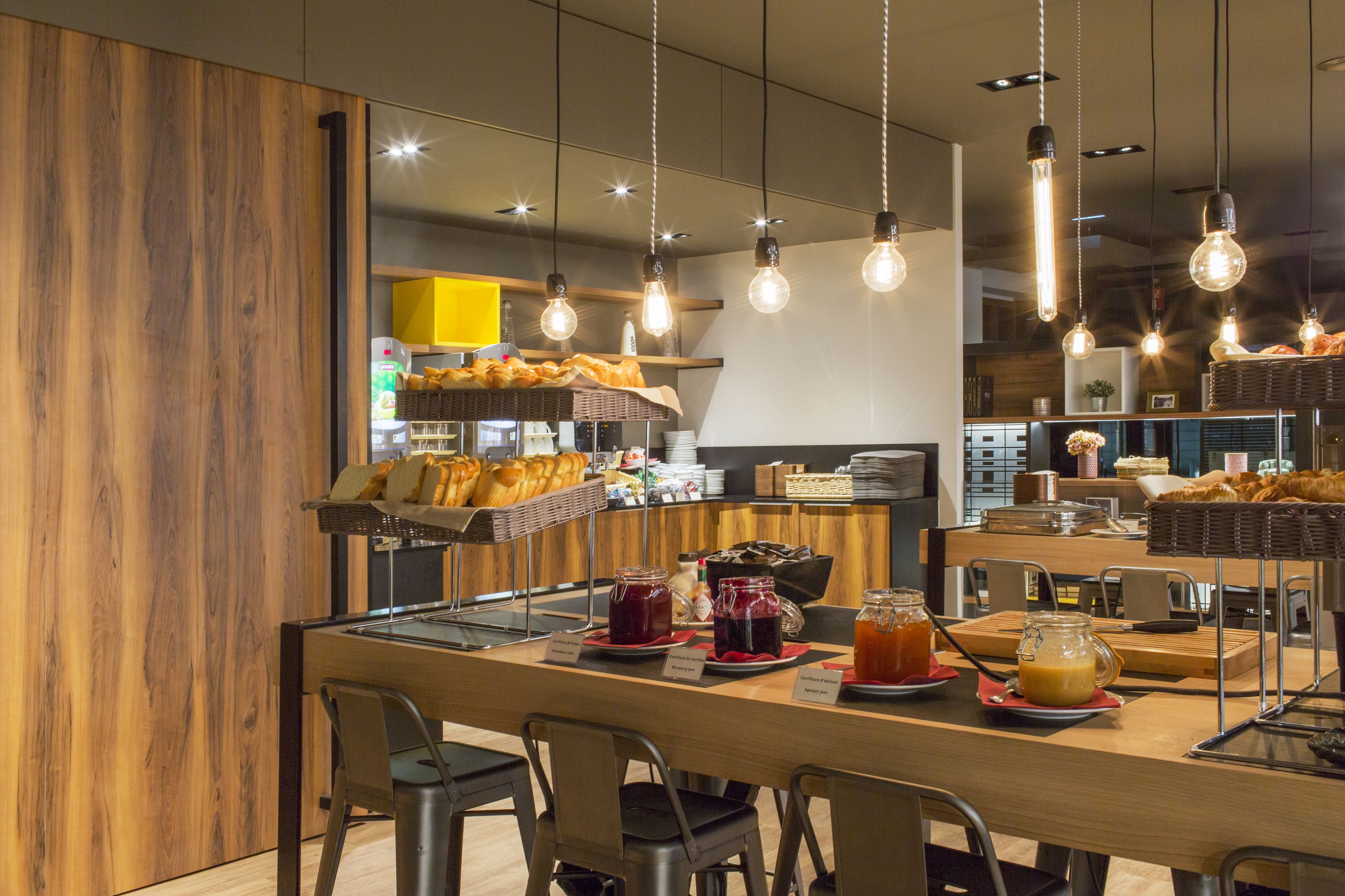 Un café moderno con mostrador de madera, luces colgantes y diversas opciones de comida y bebida.