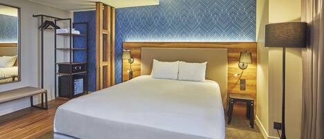 A hotel room with a large bed, a bench, a wardrobe, a desk, and a lamp.