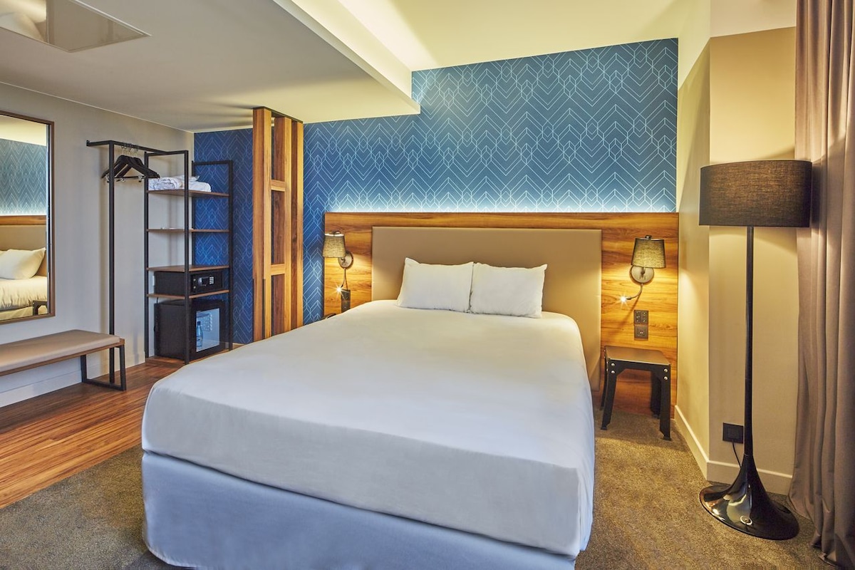 A hotel room with a large bed, bedside tables, a bench, a wardrobe, and a patterned wall.