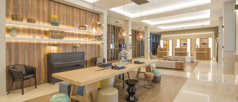 A spacious, modern hotel lobby with a wooden reception desk, comfortable seating, and a large dining table.