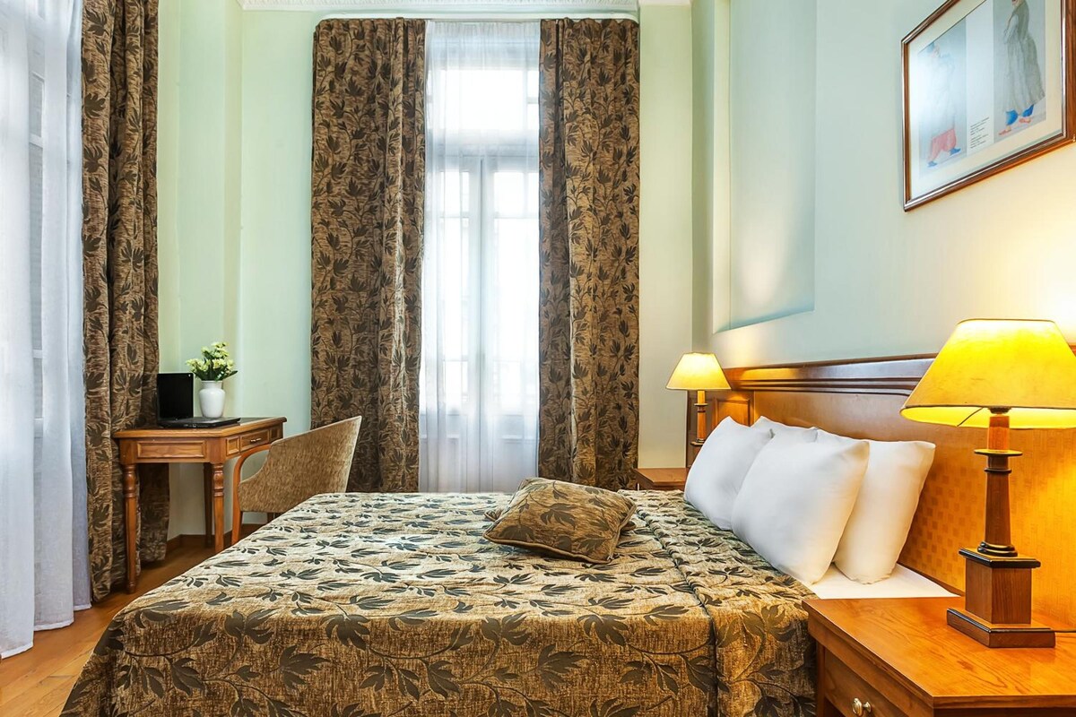 Habitación de hotel con cama, dos lámparas de noche, un escritorio con silla y una ventana con cortinas.