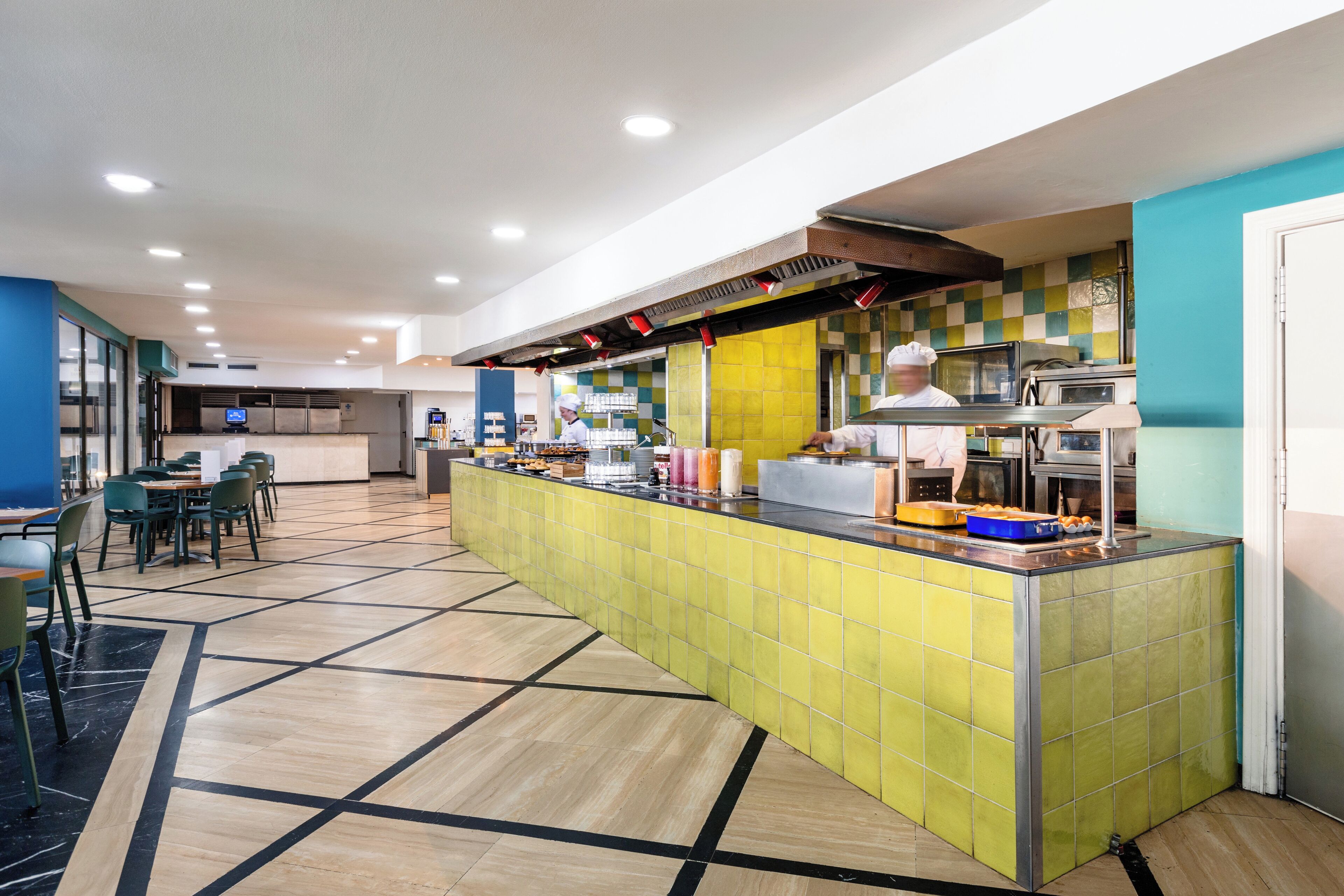 Un área de cocina moderna con una isla central, un comedor con sillas y mesas, y un chef visible al fondo.
