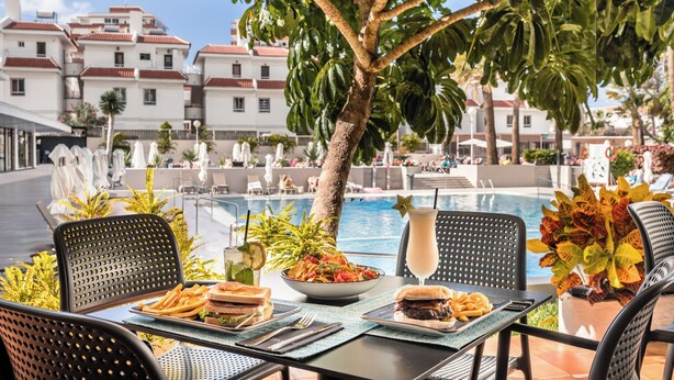 A dining table with a salad, burger, and fries, overlooking a pool with lounge chairs and umbrellas. - Catalonia Oro Negro (Arona)