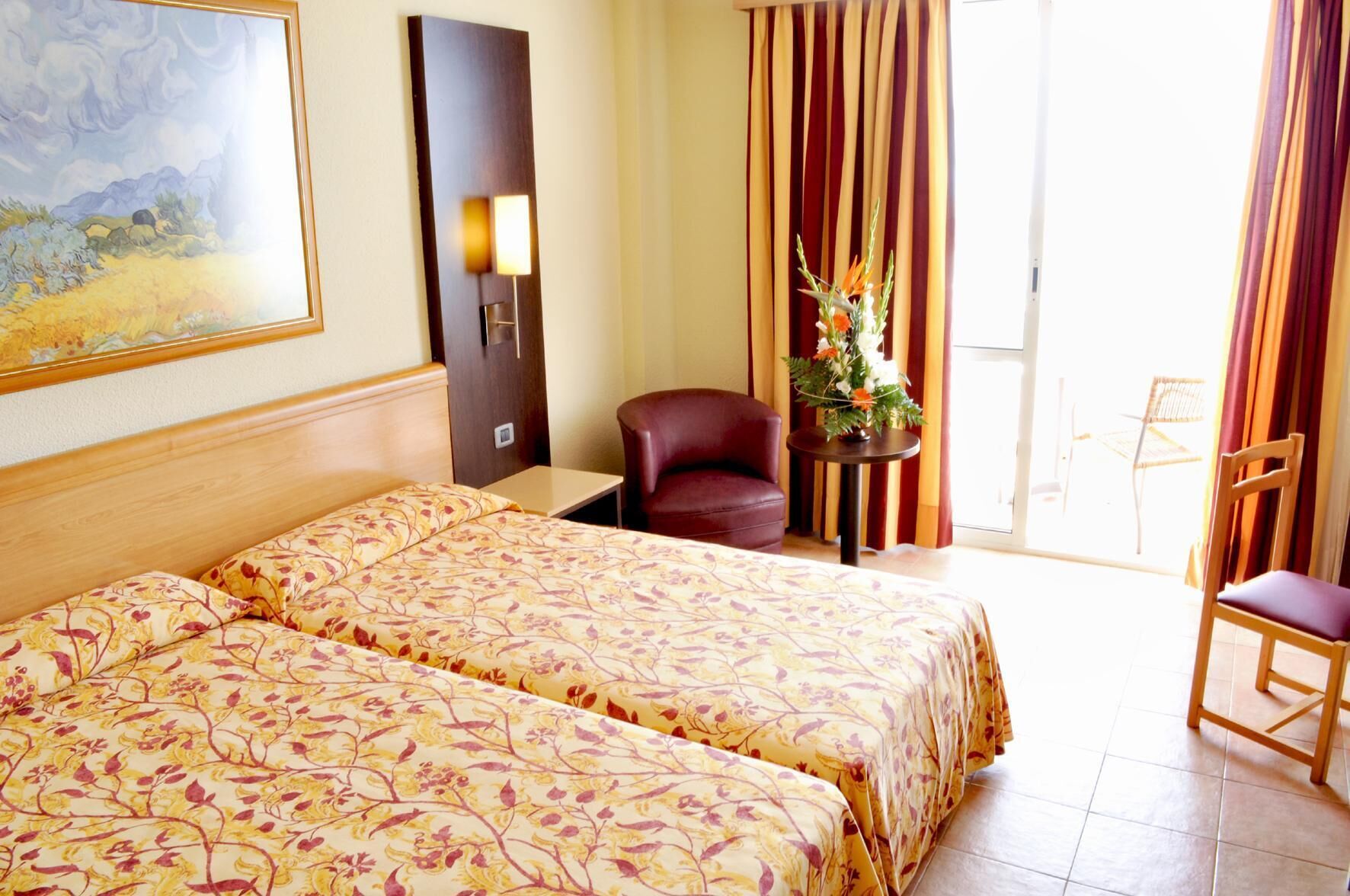 A hotel room with a bed, a chair, a desk, a lamp, and a painting on the wall.