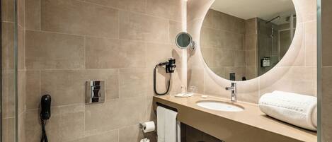 A modern bathroom with a beige tiled wall, a round mirror, a sink, a toilet, and a black stool.
