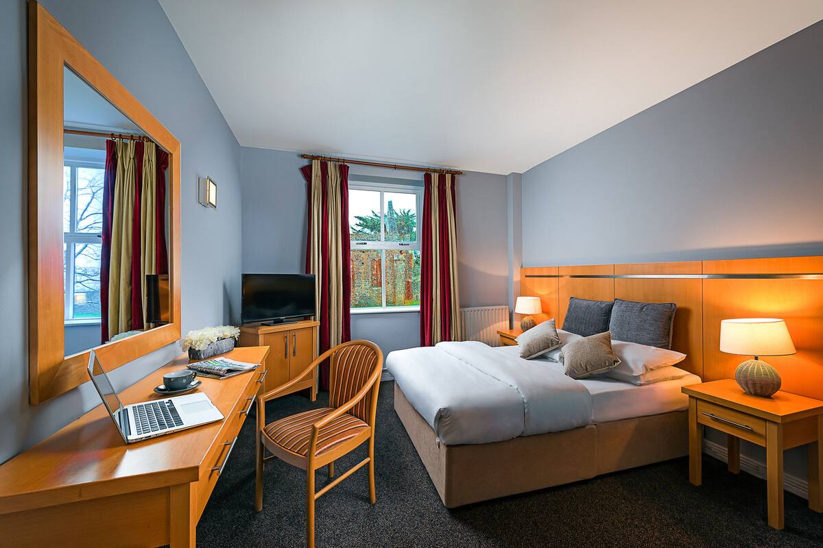 A hotel room with a bed, a desk with a laptop, a chair, a TV, and a nightstand with a lamp.
