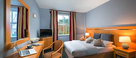A hotel room with a bed, a desk with a laptop, a chair, a TV, and a nightstand with a lamp.