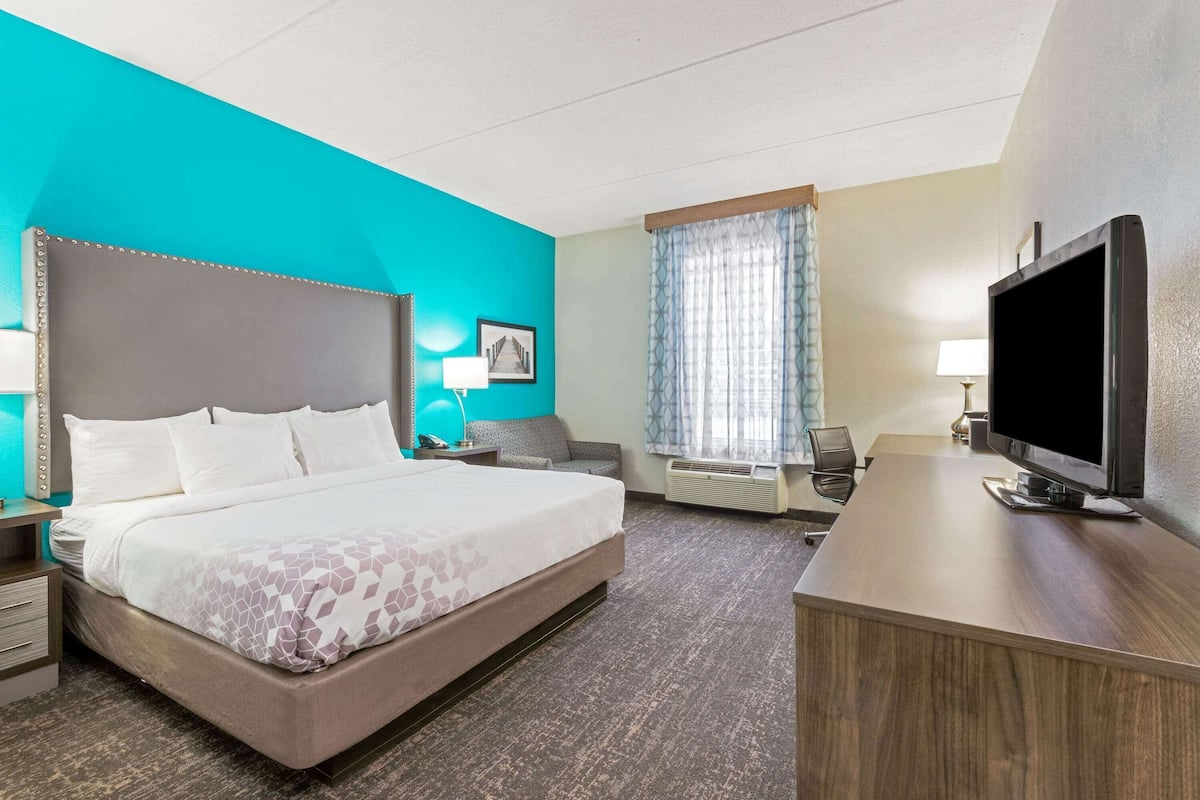 A hotel room with a large bed, a desk with a flat-screen TV, a chair, a desk lamp, and a window with curtains.