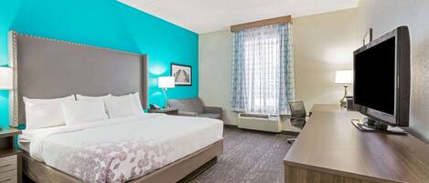 A hotel room with a large bed, a desk with a flat-screen TV, a chair, a desk lamp, and a window with curtains.