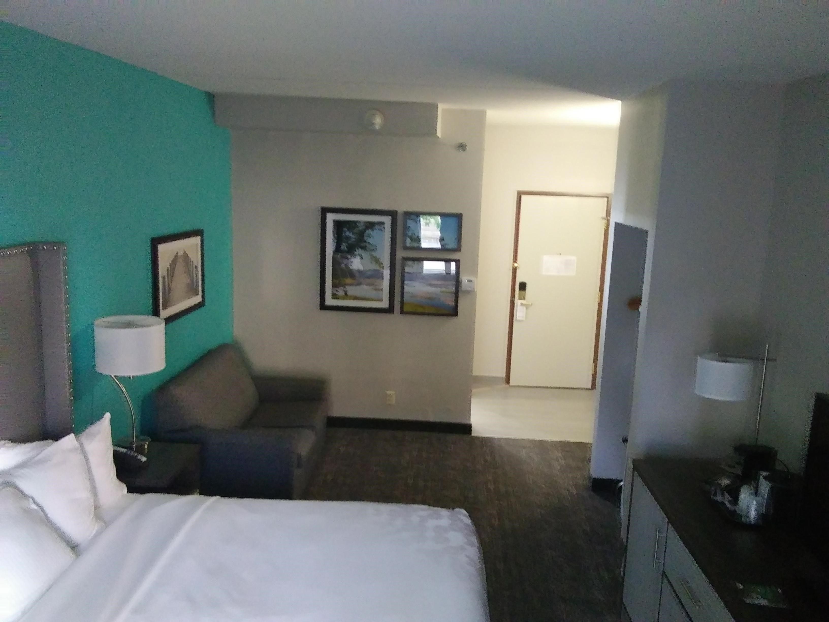A hotel room with a bed, a desk, a chair, a lamp, and a door.