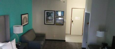 A hotel room with a bed, a desk, a chair, a lamp, and a door.