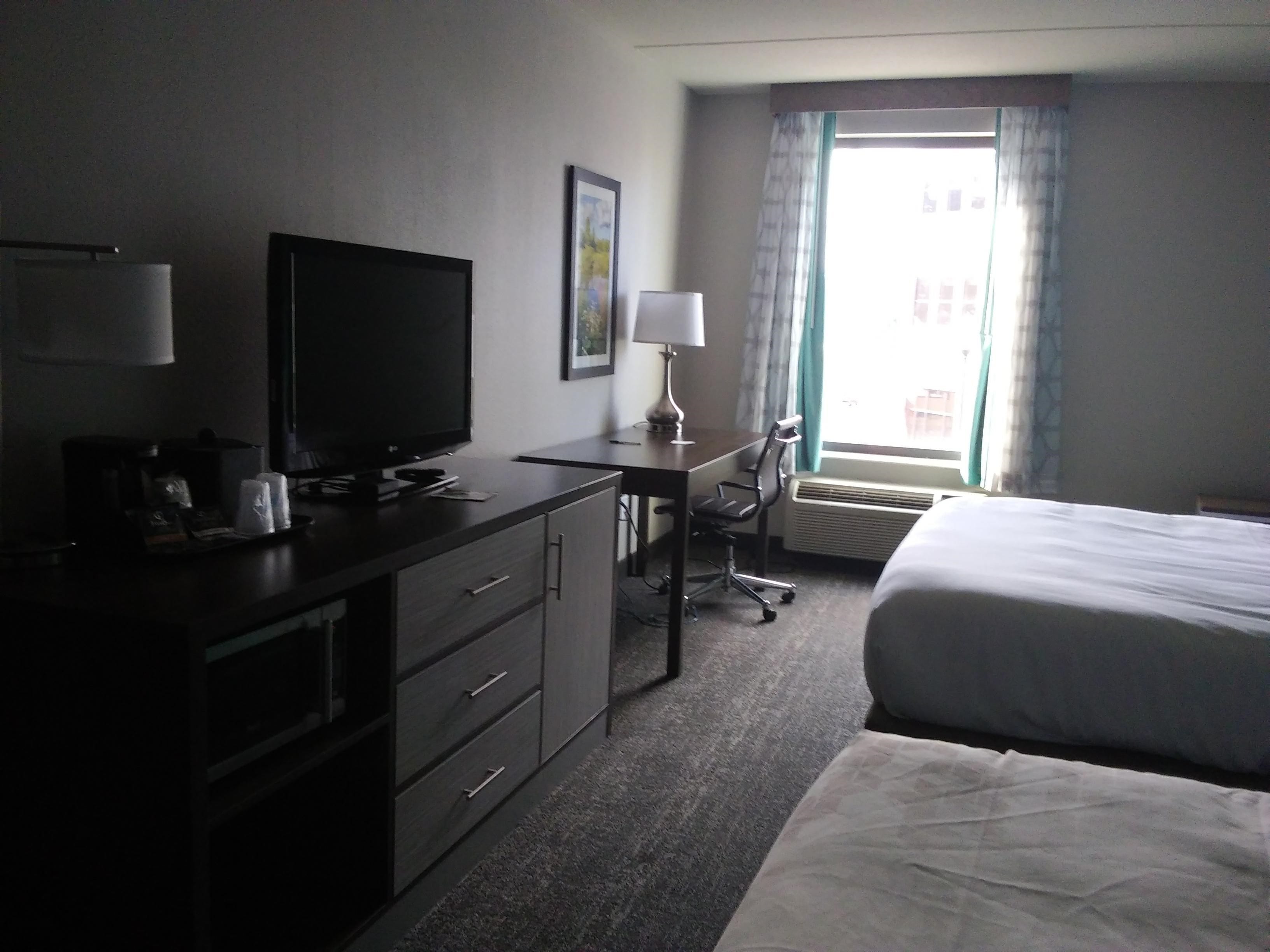 Una habitación de hotel con una cama, un escritorio con computadora, un televisor y una ventana con cortinas.