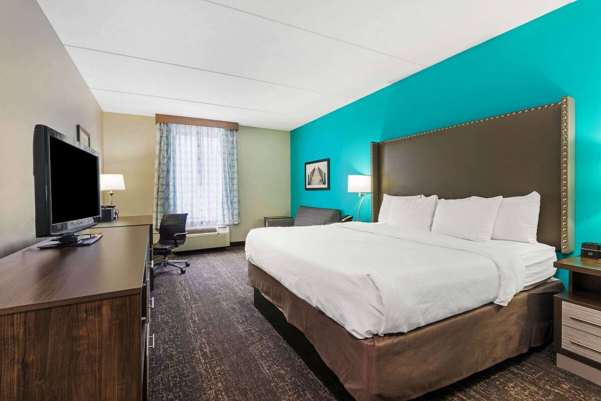 A hotel room with a large bed, a desk with a TV, a chair, a small table, and a window with curtains.